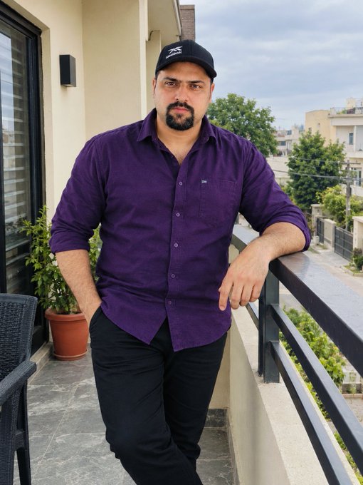 A bearded man in a purple button-up shirt and black cap leans against a balcony railing on an outdoor terrace.