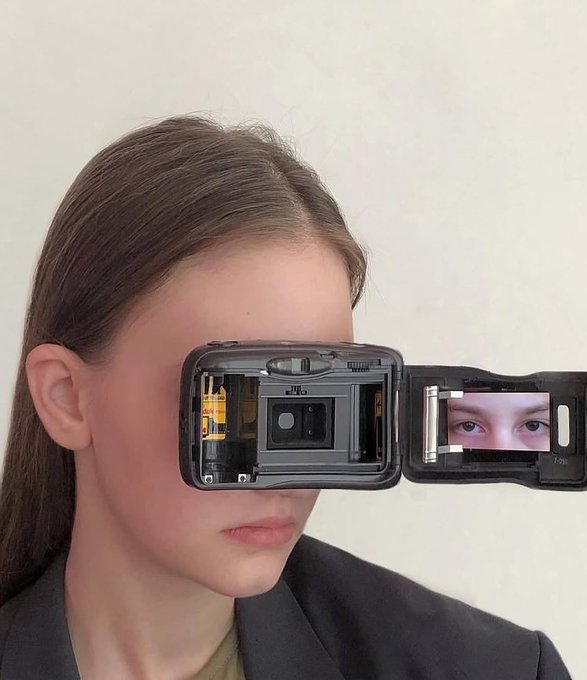 A young woman with long brown hair looks through a disassembled flip phone, its casing opened to reveal the internal components and a small screen displaying her eyes in a mirror-like reflection.