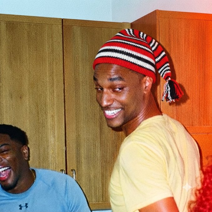 A smiling young man wearing a red, black, and white striped knit hat, with another person laughing in the background.