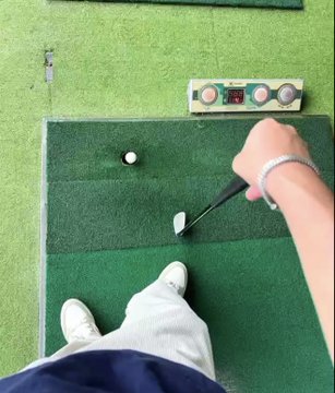 The image shows a person's feet standing on a green golf course, holding a golf club with a white ball in their hand. The person appears to be preparing for a swing or practicing their golf skills. In the background, there are two more golf clubs visible, suggesting that this is an outdoor golf practice area.