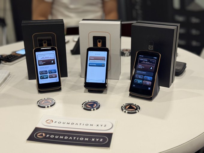 Three 'Foundation.xyz' hardware devices are displayed on a white table alongside their boxes and branded stickers, showing screens with various app interfaces.