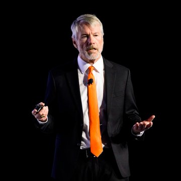 Michael Saylor, known for his pro-Bitcoin advocacy, speaks on stage in a dark suit and bright orange tie, likely during a presentation or interview promoting corporate Bitcoin adoption as a strategic move.