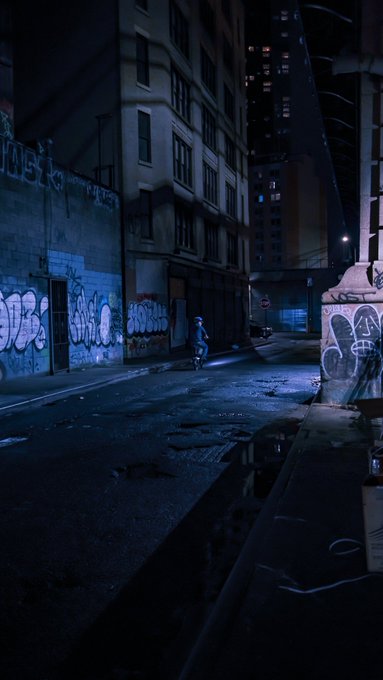 A dark, moody nighttime cityscape showing a lone figure walking down a wet alleyway with graffiti-covered walls, illuminated by cool blue light against tall buildings.