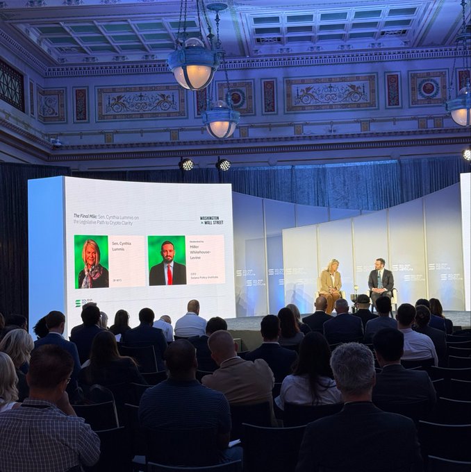 Two people sit on a stage during a panel discussion titled 'The Food Map: Sex, Crypto summit on the Legislative Path to Crypto Clarity' with speakers Sen. Cynthia Lummis and Rep. Wiley Nickel, as an audience watches from the 'WASHINGTON WALLC STREET' event.