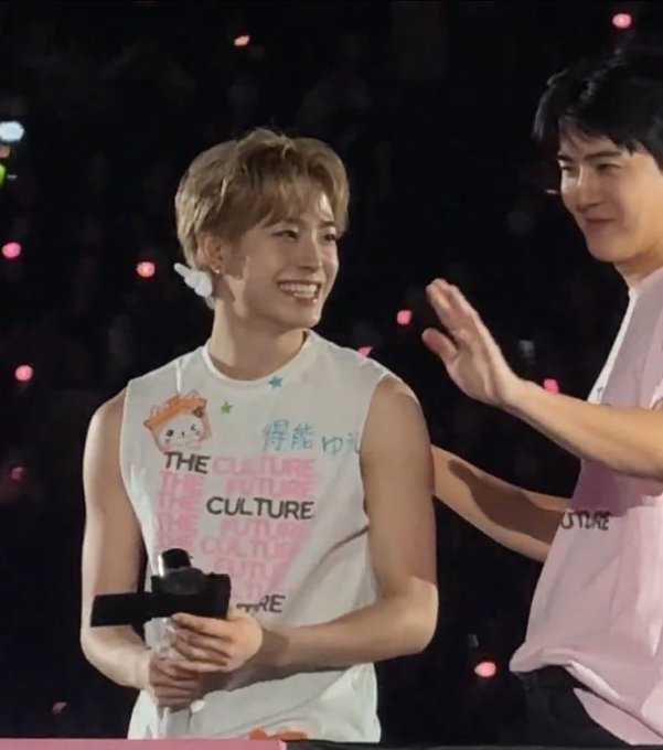 A young performer with blonde hair smiles brightly on stage, wearing a white sleeveless shirt that reads "THE CULTURE" with Asian characters and holding a microphone, while another person in a pink shirt stands beside him with a hand raised. The cheerful, intimate moment captures the joyful fan interaction the post describes at the concert's end.