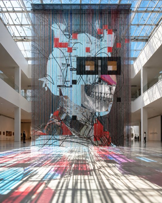 An interior photograph of a modern art gallery with a massive hanging installation depicting a stylized skull figure with red and blue tones, with colorful light patterns reflected on the floor.