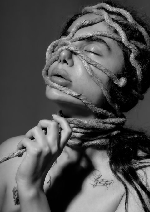 A black and white photograph captures a woman with her eyes closed, her face and neck intricately wrapped in thick rope, which she holds with one hand.