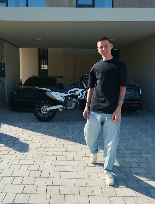 A man in a black t-shirt and jeans stands smiling in a driveway, with a black sports car and dirt bike visible in the garage behind him.