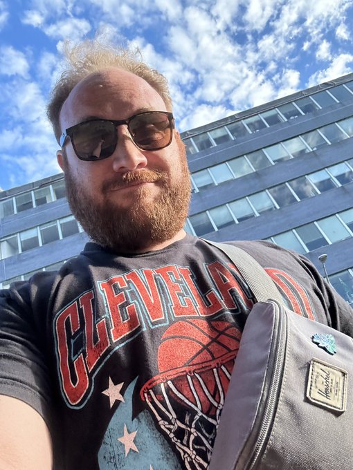 A selfie of a bearded man wearing sunglasses and a dark 'CLEVELAND' basketball graphic t-shirt, with a gray backpack strap visible and a modern glass building behind him.