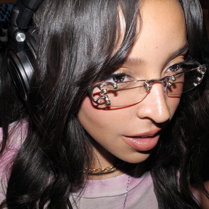 A close-up portrait shows a young woman with long dark hair wearing large headphones, a lavender top, and distinctive glasses adorned with skull charms on the temples.