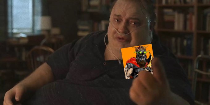 The image features a man wearing a black shirt sitting down with his hands behind him, holding up an orange card that has a picture of a person with large eyes and a beard printed on it. The caption reads "It's Father's Day and Bitcoin Cyborgs are fatherless orphans". The overall setting appears to be indoors in a room filled with bookshelves where multiple books can be seen.