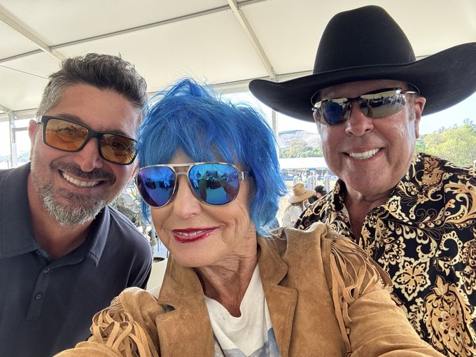 Three people pose for a selfie under a white canopy; a man on the left wears yellow-tinted glasses, an older woman in the middle sports vibrant blue hair and reflective aviator sunglasses, and a man on the right wears a black cowboy hat, sunglasses, and a black-and-gold patterned shirt.