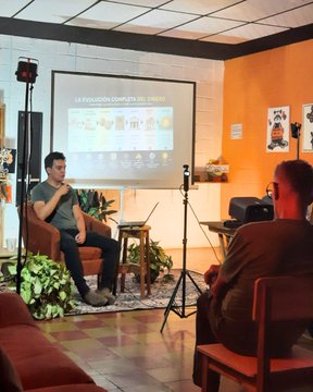 A man sits in an armchair speaking to an audience in an orange-walled room, with a projector screen behind him displaying a presentation titled 'LA EVOLUCIÓN COMPLETA DEL DINERO'.
