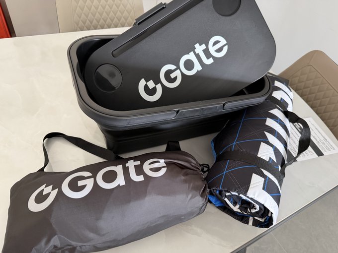 A black hard-shell case branded "Gate" sits open on a marble table alongside a matching drawstring bag and a rolled-up patterned picnic mat, showcasing the camping merchandise gifted by Gate for Mid-Autumn Festival.