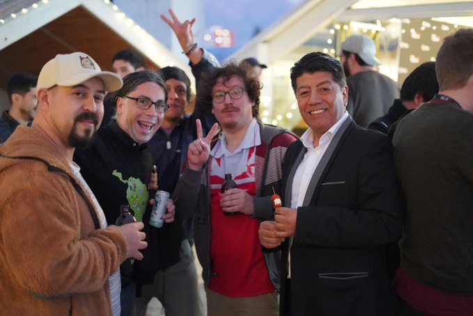 A group of four men smiling and posing at an outdoor evening event, with one wearing a red Arsenal jersey and another in a brown jacket, with tents and string lights in the background.