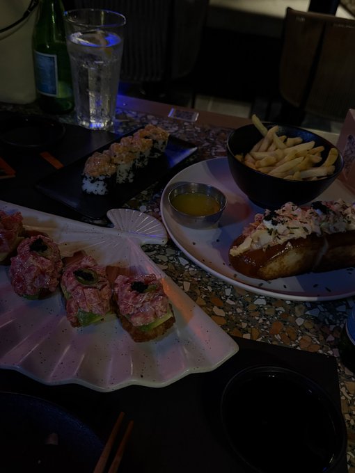 A dimly lit table at a restaurant features a spread of food including sushi rolls topped with what appears to be tuna tartare, a bowl of french fries, bruschetta with toppings, and a small dish of sauce, accompanied by a glass of water and a green bottle.