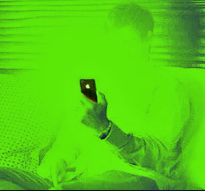 A person holds a glowing smartphone in a room completely bathed in intense green light, obscuring the surrounding details.