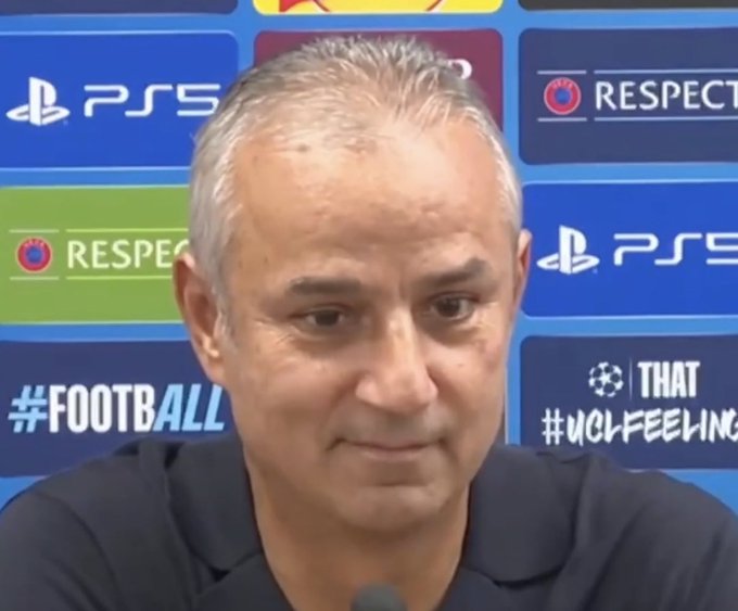 A grey-haired man sits at a press conference in front of a backdrop featuring "PS5," "RESPECT," and "#FOOTBALL #UCLFEELINGS" logos, appearing serious as he addresses media. The image contrasts with the accompanying crypto-trading brag post, highlighting how online self-promotional content about Bitcoin and altcoins diverges from mundane real-world moments like a UEFA Champions League press briefing.
