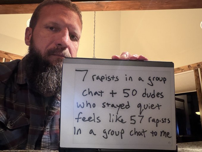 A bearded man holding up a whiteboard that reads, "7 rapists in a group chat + 50 dudes who stayed quiet feels like 57 rapists in a group chat to me".