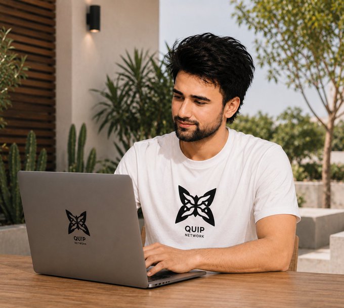 A man with dark hair and a beard sits outdoors at a wooden table working on a laptop, wearing a white t-shirt featuring a black geometric logo and the text 'QUIP NETWORK'.