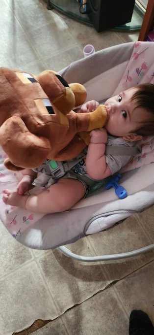 The image features a baby lying down in a white rocking chair, with a large brown teddy bear next to them. A caption below the photo reads "He loves his @doginaldogs", which seems to be related to the teddy bear or the baby's name. The presence of the teddy bear and the baby's name suggests that this image might have been taken during a special occasion, such as a birthday party or a family gathering where the baby is being celebrated with a cute and cuddly stuffed animal companion.