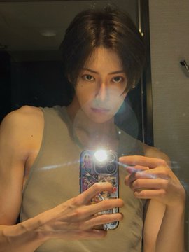 A young man with long dark hair takes a mirror selfie wearing a tan tank top, with a bright camera flash partially obscuring his face. The hashtags reference Alpha Drive One (ALD1), the K-pop project group featuring the member ARNO, matching the person's appearance.