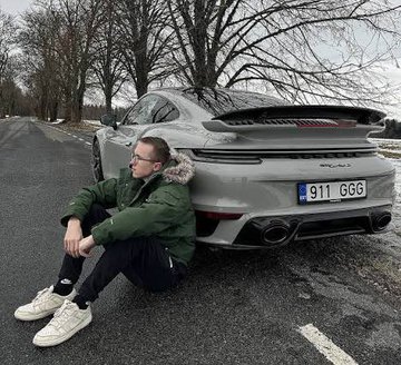 A man sits on the ground next to a silver Porsche 911 GT3 RS with the license plate "911 GGG" parked on a rural road. The image illustrates the Polymarket post about professional CS:GO player ropz attending an EWC event in Paris with his Porsche, with a 13% betting chance he'll get pulled over again like his previous incident.