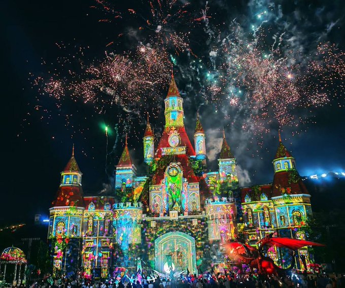 A vibrant nighttime scene of a castle-like building elaborately decorated with colorful projected imagery and projections of Disney characters, with spectacular fireworks exploding in the sky above and a crowd gathered in front.