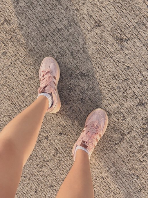A first-person view looking down at dusty pink running shoes on a textured pavement, perfectly answering the flash task's request for a running shoes photo. The legs are stretched out ahead, suggesting the wearer is mid-stride or paused during a run — a casual, on-the-go fit for the $INJ squad challenge.