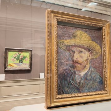 A museum display shows a large framed self-portrait of a bearded man in a straw hat next to a smaller framed sunflower painting.