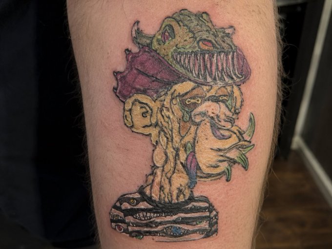 A tattoo on skin depicting a monstrous creature with sharp teeth, fangs, and claws perched atop a stacked pile of books.