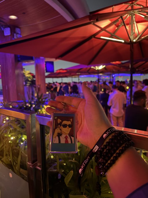 A first-person view of a hand holding up an event badge featuring an anime-style character with sunglasses and a red tie, taken at an outdoor evening gathering with red umbrellas and Kraken wristbands visible.