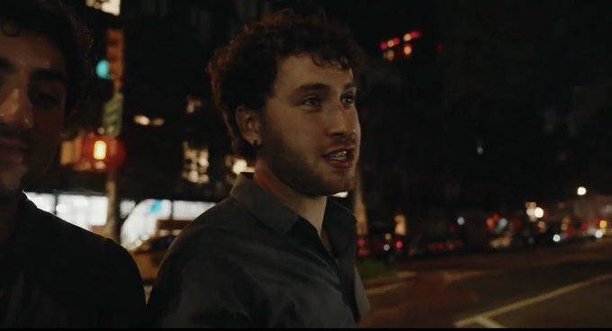 A nighttime shot of a young man with curly hair and a beard, wearing a dark collared shirt, speaking while standing on a city street with illuminated buildings and traffic in the background.