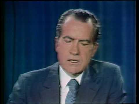 A vintage color photograph of a man in a suit and tie against a blue curtain backdrop, appearing to be from a historical television broadcast.