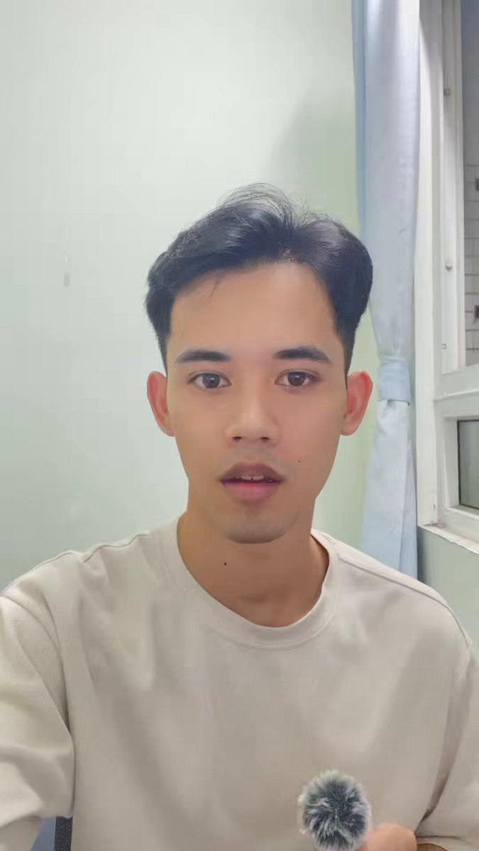 The image features a young man with short hair wearing a white shirt who appears to be taking a selfie or making a video call using his phone's camera. The background of the photo shows a window, suggesting that he might be indoors or near an outdoor area like a balcony. The man is holding a microphone in one hand and a small object in the other, possibly related to the content being shared on social media platforms.