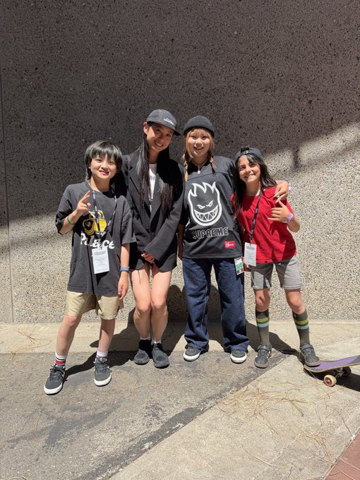 A group photo of four young people posing together outdoors against a gray wall, with one wearing a Supreme hoodie and another in a red shirt near a skateboard.