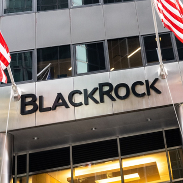 The image shows the exterior of a BlackRock office building entrance, featuring the company's name prominently displayed in bold black lettering on a silver facade, flanked by American flags. This visual ties to the news about BlackRock's recent decision to lower the minimum for in-kind transfers to $1 million for its Bitcoin ETF, making the fund more accessible to institutional investors.