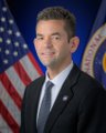 The image shows Jared Isaacman, identified by his official NASA Administrator portrait pose with an American flag and NASA seal in the background, visually connecting him to the post discussing his announcements about NASA's Artemis program timeline and the Dragonfly mission.