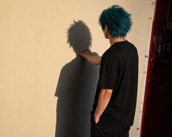 A person with blue hair wearing a black t-shirt stands facing a beige wall, reaching out to touch their dark shadow cast upon it.