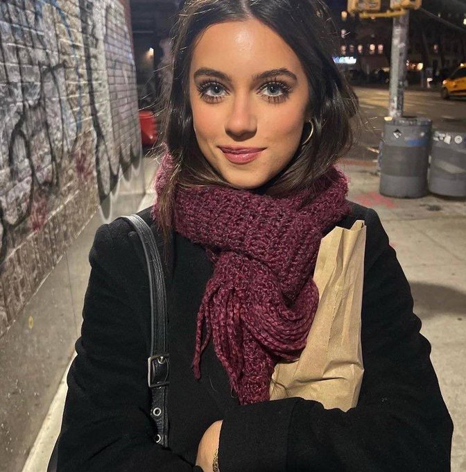 A nighttime photo of a young woman with brown hair wearing a black coat and burgundy knit scarf, standing on a city sidewalk near trash cans and a graffiti-covered wall.