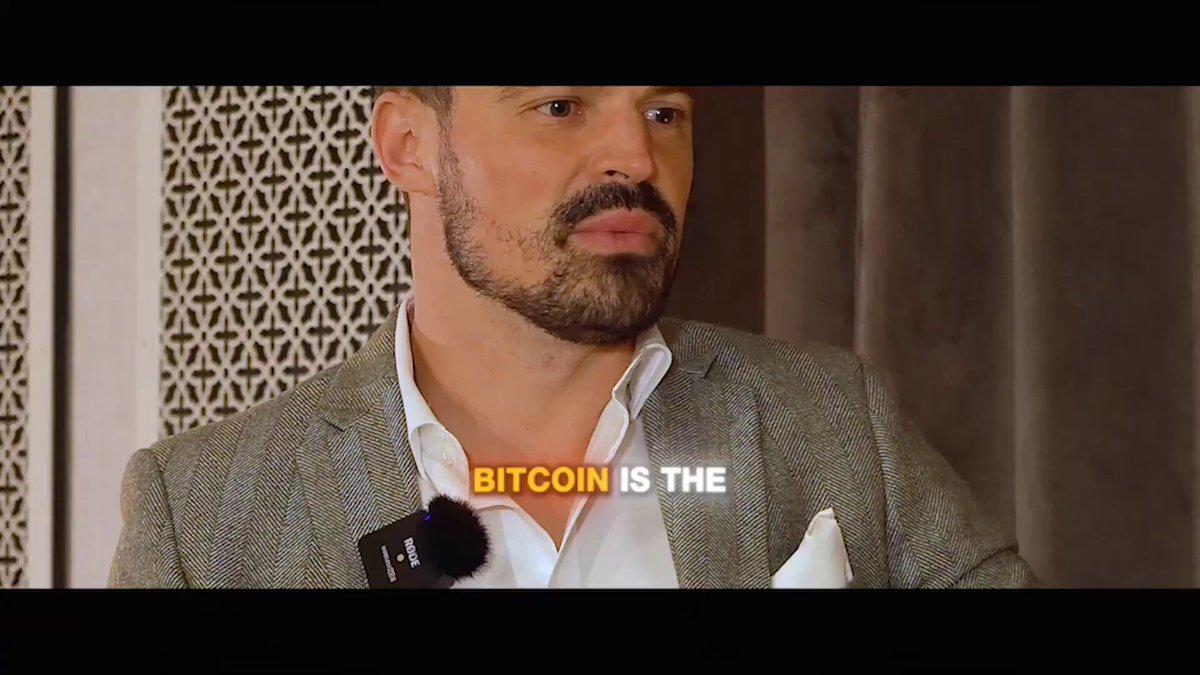 The image shows a man wearing a suit and tie standing against a gray background with white text that reads "BITCOIN IS THE". The man appears to be speaking into a microphone or recording device, possibly at an event like the Income Show where they are discussing the future of money, technology, digital credit, AI, and Bitcoin.