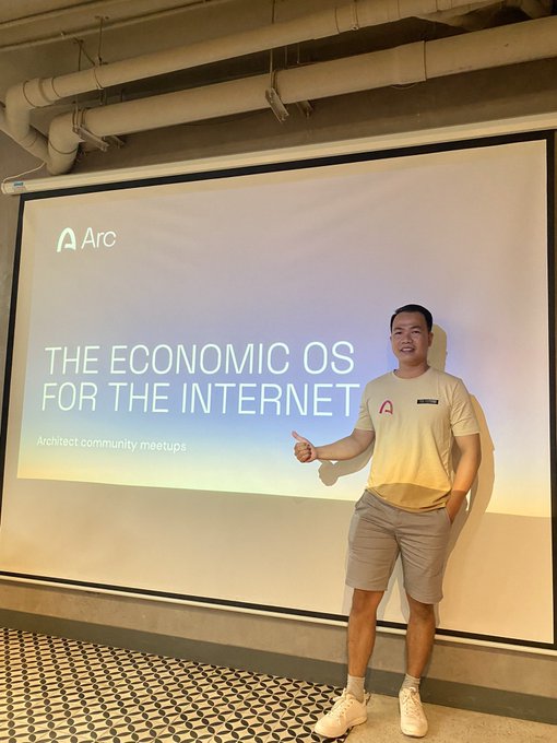 A presenter wearing an "Arc" branded t-shirt stands smiling in front of a large projection screen displaying "THE ECONOMIC OS FOR THE INTERNET" along with the Arc logo and "Architect community meetups" text, illustrating the speaker delivering a talk at the Arc meetup held in Ho Chi Minh City, Vietnam.
