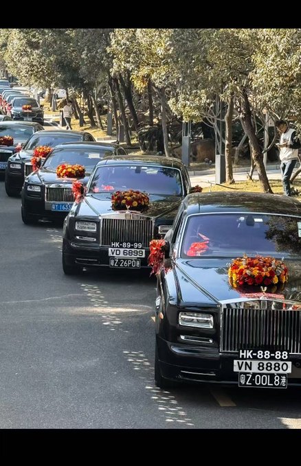 A wedding procession featuring a line of black Rolls-Royce cars decorated with red floral arrangements on tree-lined streets.
