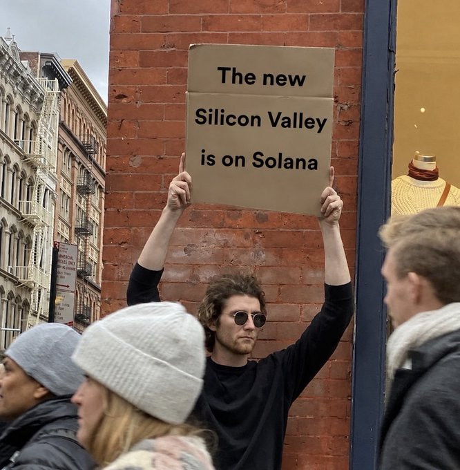 A man with sunglasses and a beard holds up a cardboard sign reading 'The new Silicon Valley is on Solana' in front of a brick building while crowds gather nearby.