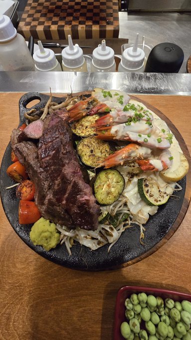 A delicious-looking surf and turf platter features a well-seared steak, succulent shrimp topped with creamy sauce and chives, grilled zucchini, charred tomatoes, bean sprouts, and a dollop of wasabi, served on a hot stone platter with squeeze bottles and edamame on the side. Despite the caption's mention of NFT trading losses from rug pulls draining the wallet, the image showcases an indulgent, comforting meal — a reminder that even after financial setbacks in the crypto space, good food remains a small, accessible pleasure to lift the spirits during a weekend night.