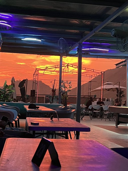 An outdoor covered patio or restaurant area at sunset, featuring tables, chairs, blue LED lights on the ceiling, and a view of an orange and yellow sky with mountains in the distance.