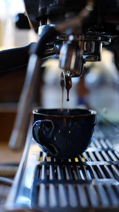 A close-up captures a stream of espresso being extracted from a gleaming chrome machine into a dark ceramic cup, resting on a slatted metal drip tray.