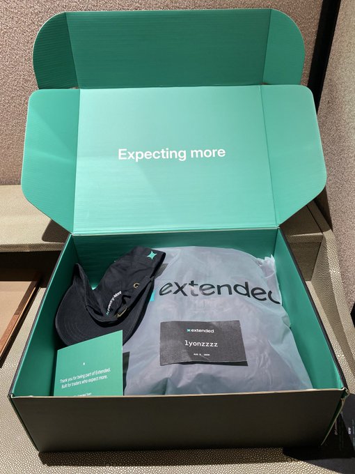 An open green Extended Stay subscription box containing a dark baseball cap and clothing items, with the inside lid reading 'Expecting more' and a label addressed to 'lyonzzzz'.