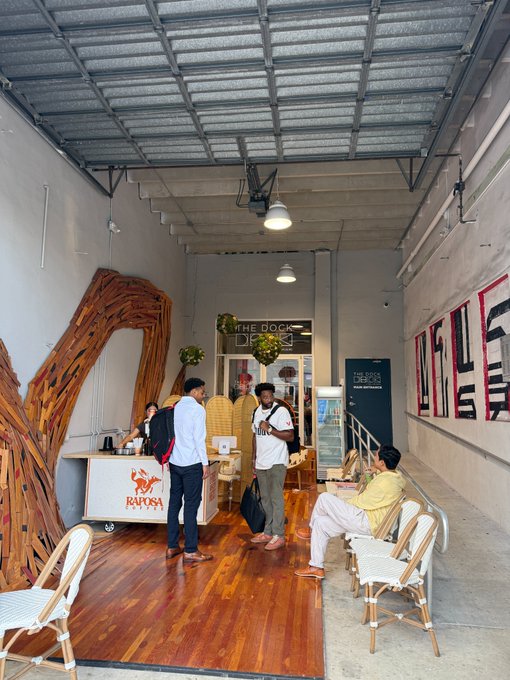 Inside a bright, industrial-style corridor, visitors gather at the "Raposa" coffee pop-up cart located just outside "The Dock," with a large "RAPOSA" banner on the wall and passersby sampling drinks while chatting in the wooden-floored space.