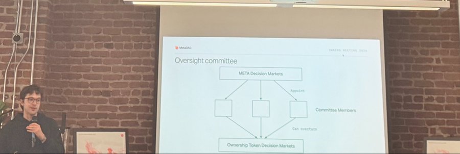 A man with glasses and dark hair holding a microphone stands in front of a projection screen displaying a diagram titled 'Oversight committee' that shows 'META Decision Markets' and 'Ownership Token Decision Markets'.